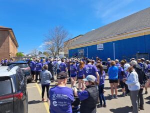 Group of people in purple Hike for Hospice t-shirts gathering outside a blue building under a bright blue sky