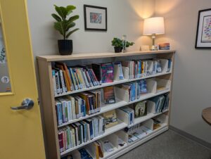 A double shelf unit, 4 shelves high, sits in the corner of the Grief Library. A yellow door opens toward the shelf. On top of the shelf are plants, a trophy, and a lamp. There are pictures on the walls.